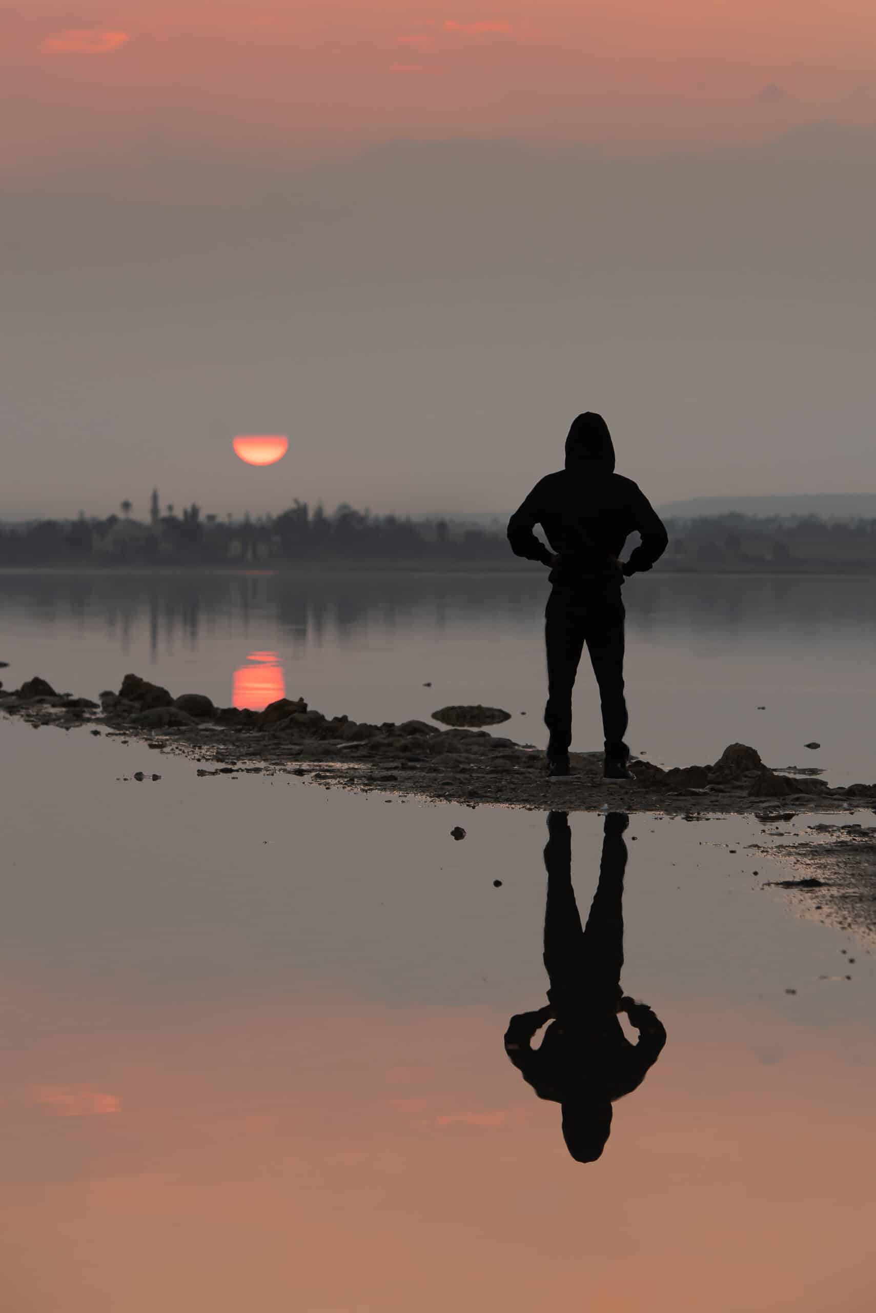 A person standing by the water at sunrise, symbolizing inner reflection, energetic symmetry, and the holistic wellness journey.
